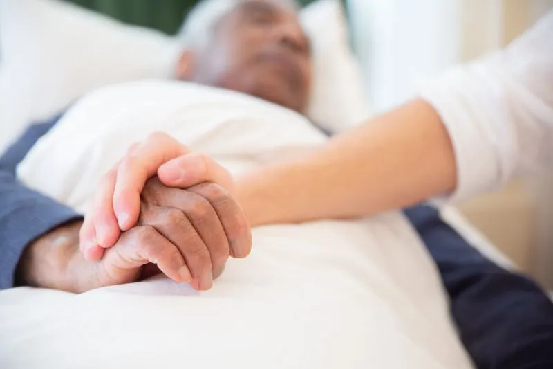 Jongere hand houdt zorgzaam de hand vast van een oudere die in bed ligt
