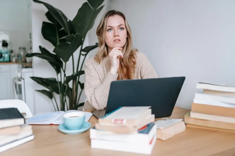 Vrouw denkt na achter haar laptop, omringd door boeken en een kop koffie